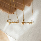 Two gold Summer Name Necklaces with a script font rest on textured paper; one says Sarah, the other Ananda—ideal as a personalized necklace set.