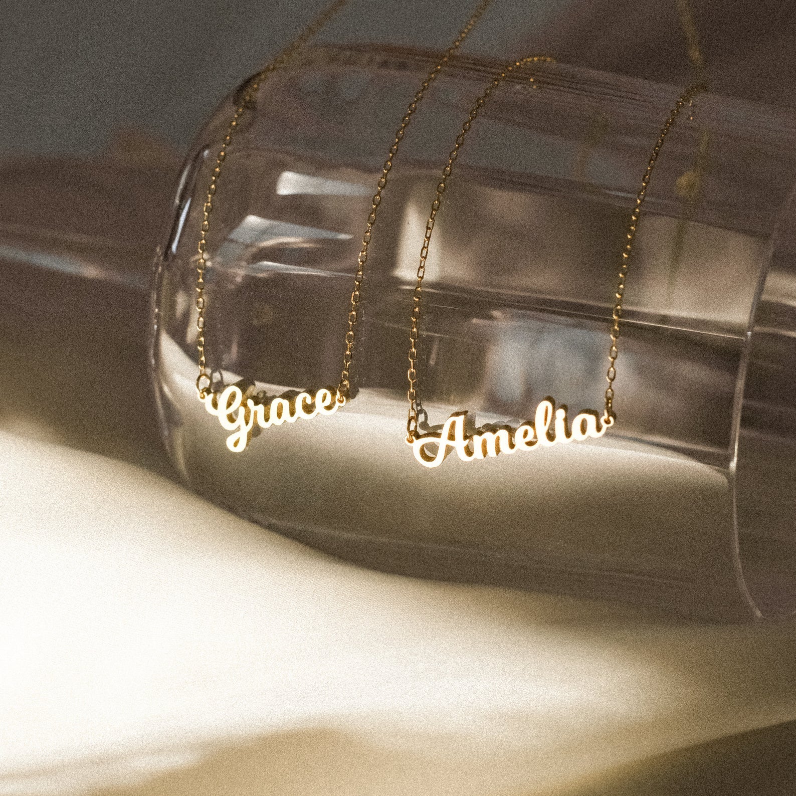 Two rose gold Mellow Name Necklaces with the names Grace and Amelia drape elegantly over a glass—a stunning showcase of the Mellow Name Necklace in Rose Gold.