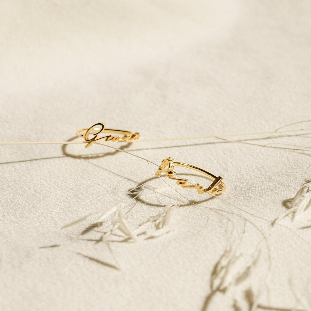 Two delicate Venice Name Rings with elegant cursive script rest on a textured cream surface, accented by dried plant stems.