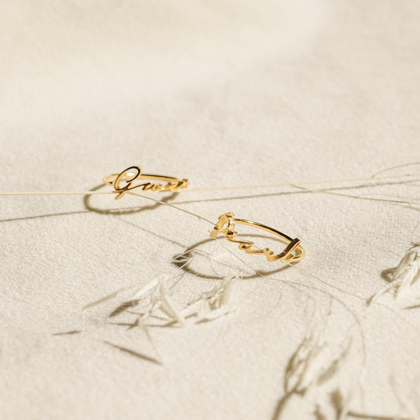 Two delicate Venice Name Rings with elegant cursive script rest on a textured cream surface, accented by dried plant stems.