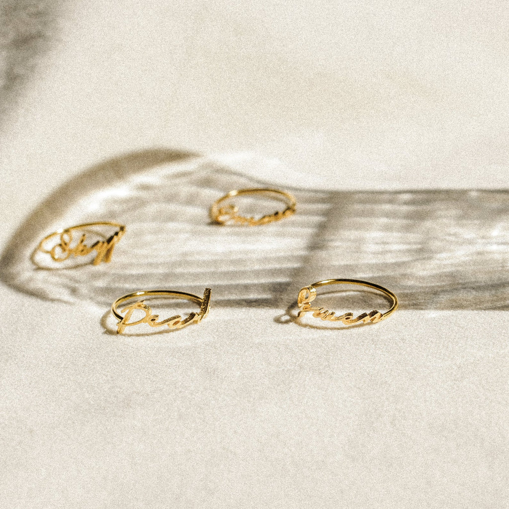 Four delicate Venice Name Rings with cursive words rest on a light surface, casting soft shadows—a beautiful touch of custom jewelry.