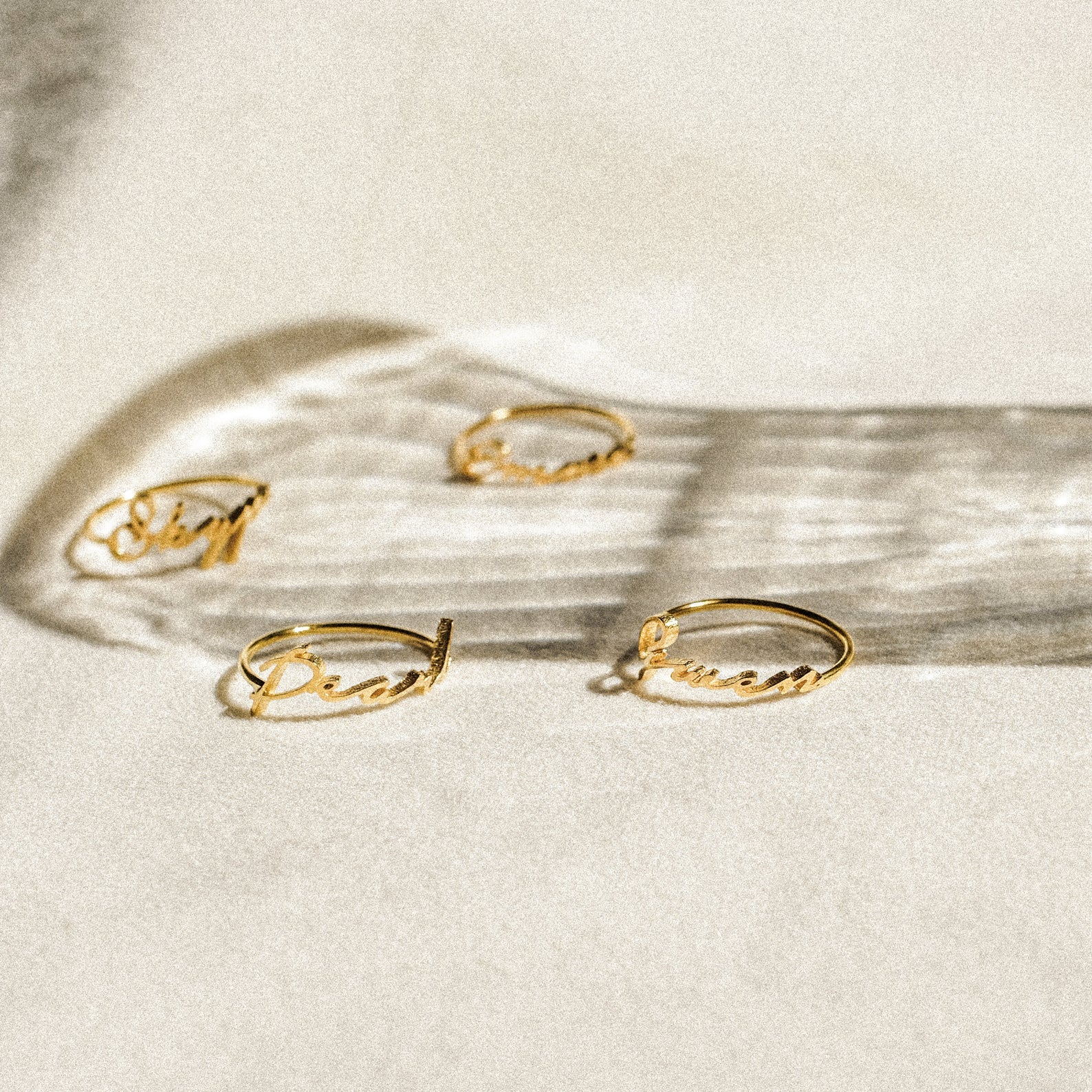 Four delicate Venice Name Rings with cursive words rest on a light surface, casting soft shadows—a beautiful touch of custom jewelry.