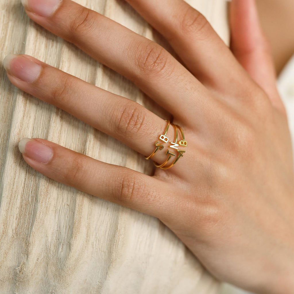 A hand with manicured nails showcases the Modern Duo Initial Ring—an elegant gold ring with minimalist, abstract accents—worn on the middle finger to highlight the sophistication of custom jewelry.