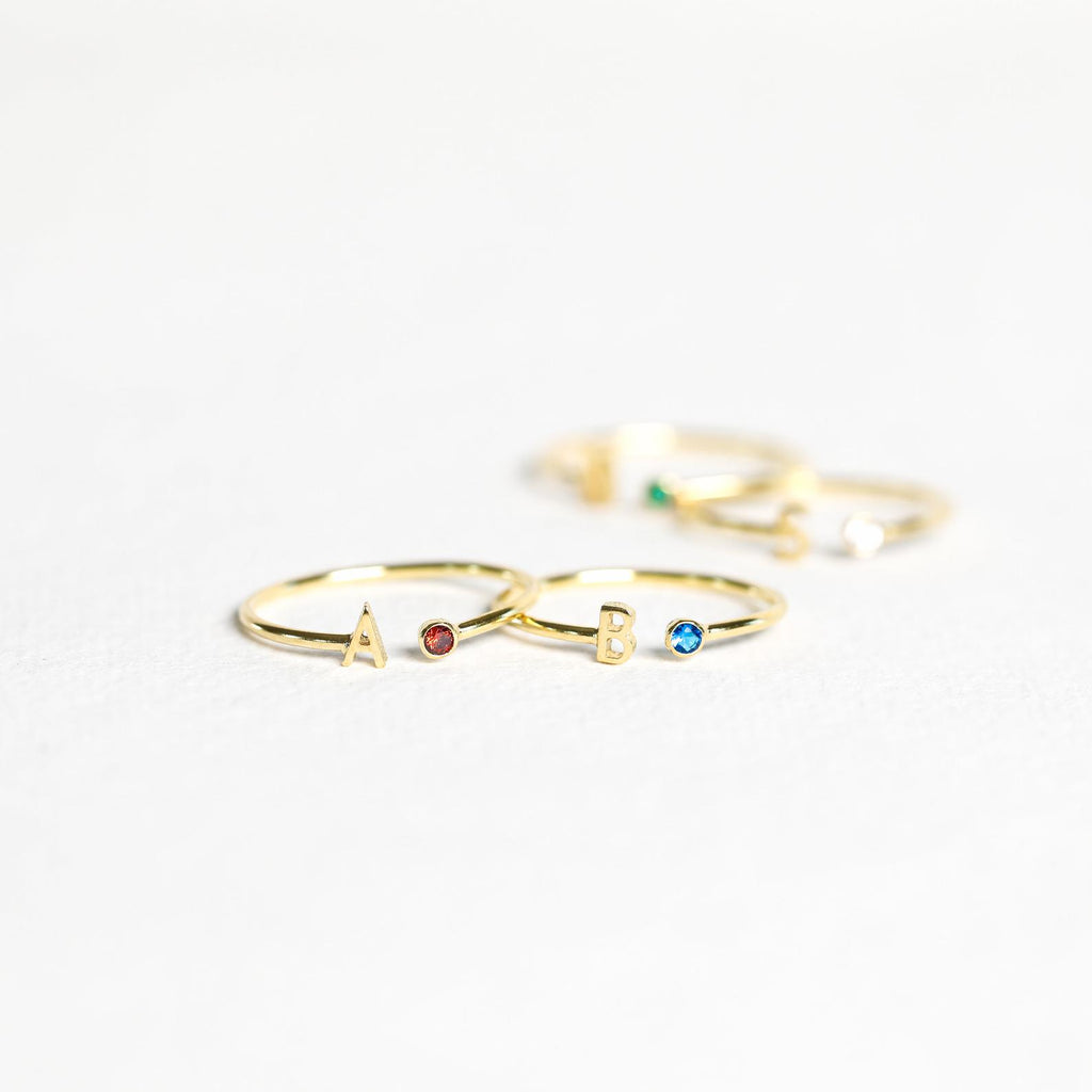 Two gold Modern Initial Birthstone Rings, each featuring an initial and a small gemstone, sit on a white surface—an ideal personalized gift for Christmas.