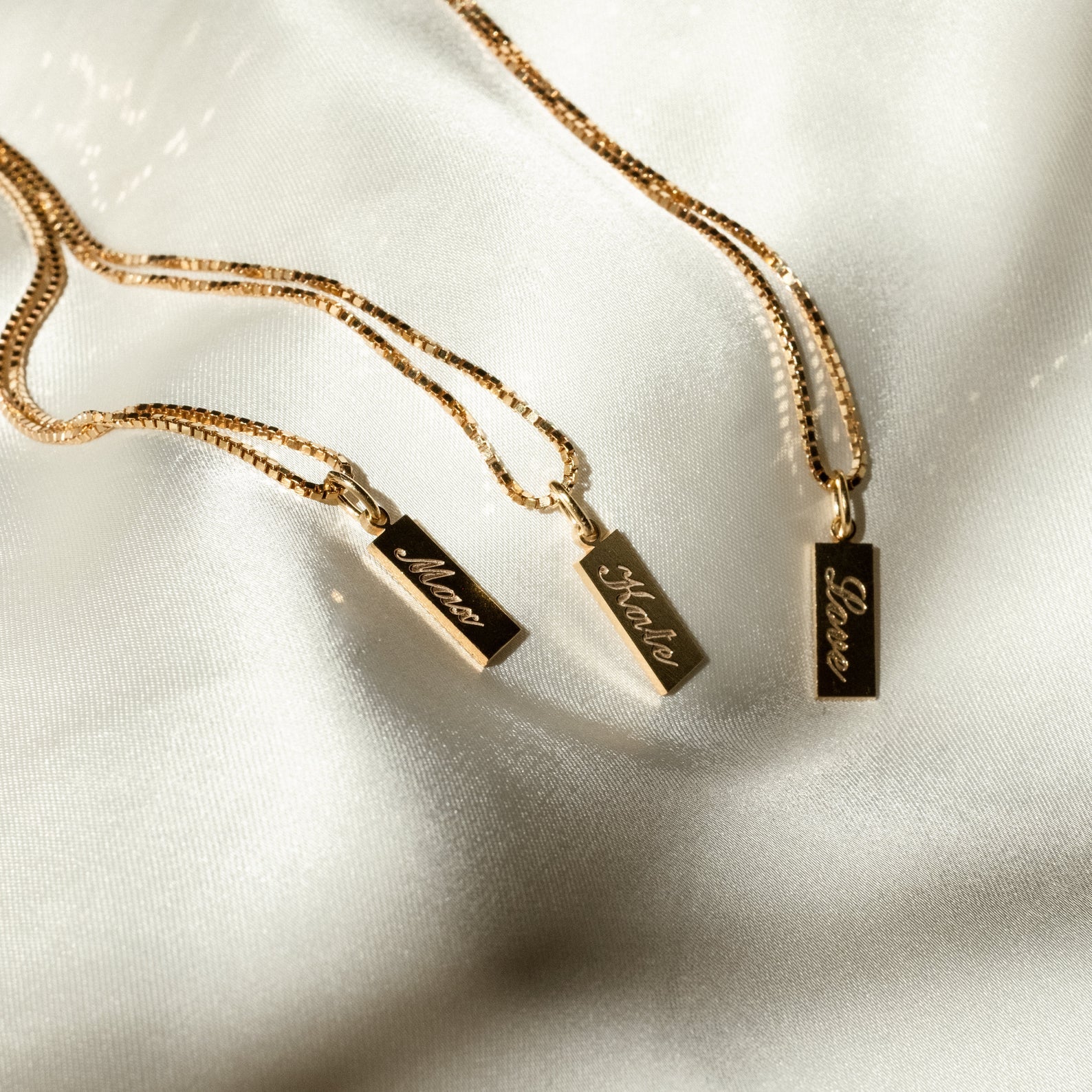 Three rose gold Dainty Tag Name Necklaces with rectangular pendants labeled Mamá, Mother, and Love rest on white satin fabric. Each features a delicate dainty chain for an elegant touch.