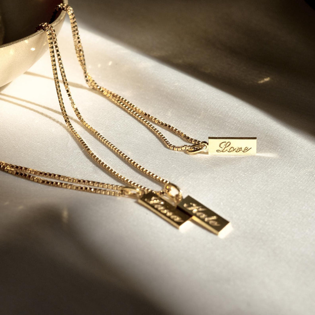 The Dainty Tag Name Necklace in Rose Gold features engraved rectangular tags reading "Love," "Faith," and "Hope" on delicate chains, creating an elegant and meaningful accessory collection displayed on a white surface.