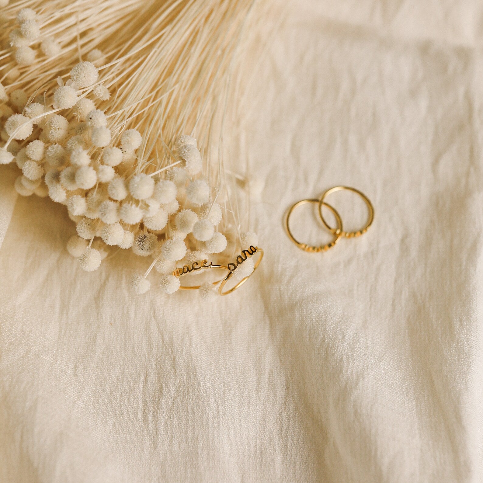 The Petite Tiny Name Ring is displayed with gold rings and dried white flowers on a cream-colored fabric background.