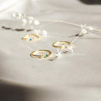 The Venice Initial Birthstone Ring, a delicate gold ring adorned with a personalized initial and colorful birthstones, is displayed on light fabric next to white dried flowers.