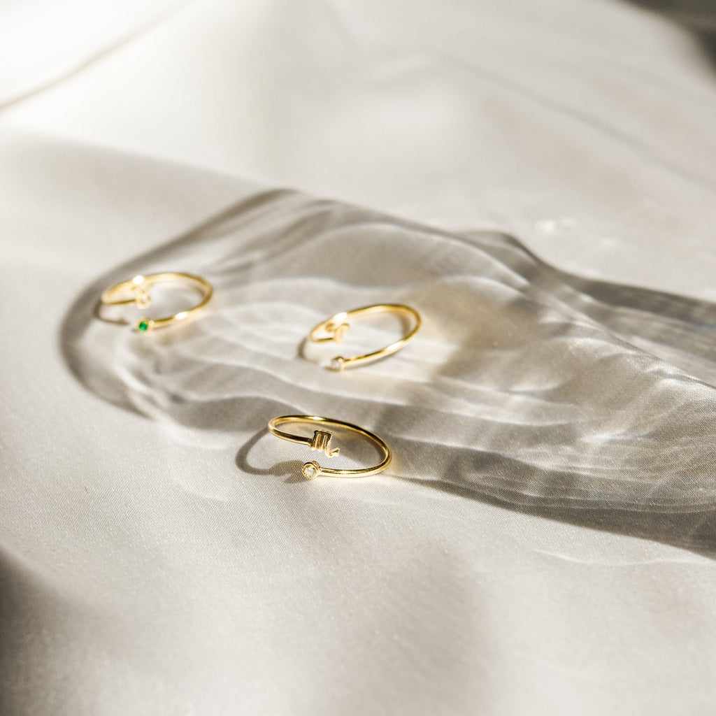 The Zodiac Birthstone Ring, a delicate gold ring adorned with a small gemstone, rests on white satin and casts soft shadows—an elegant piece ideal as a zodiac or birthstone ring.