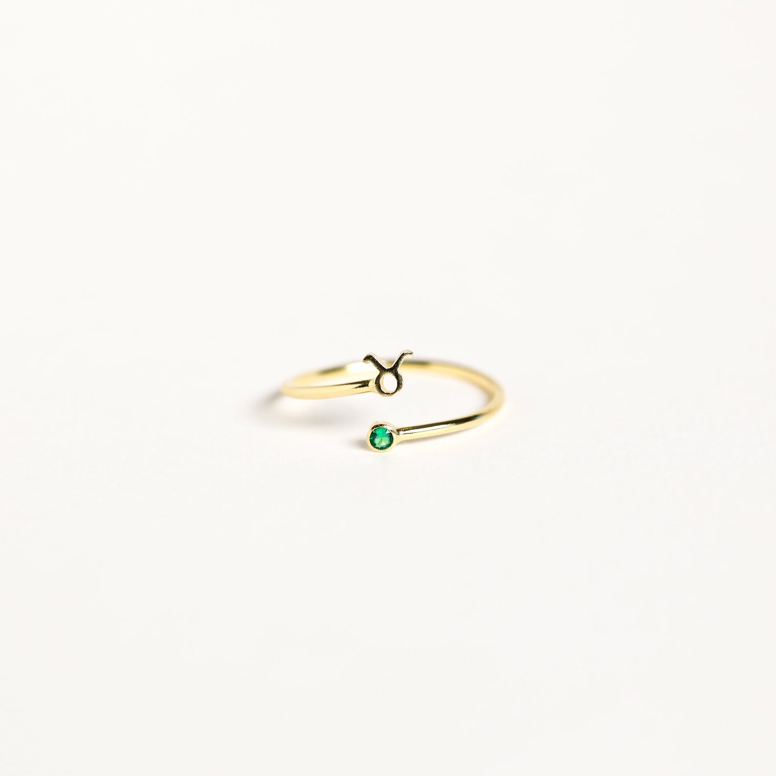 The Zodiac Birthstone Ring features a gold band with the Taurus symbol and a small green birthstone, combining elegant jewelry with celestial charm on a plain white background.