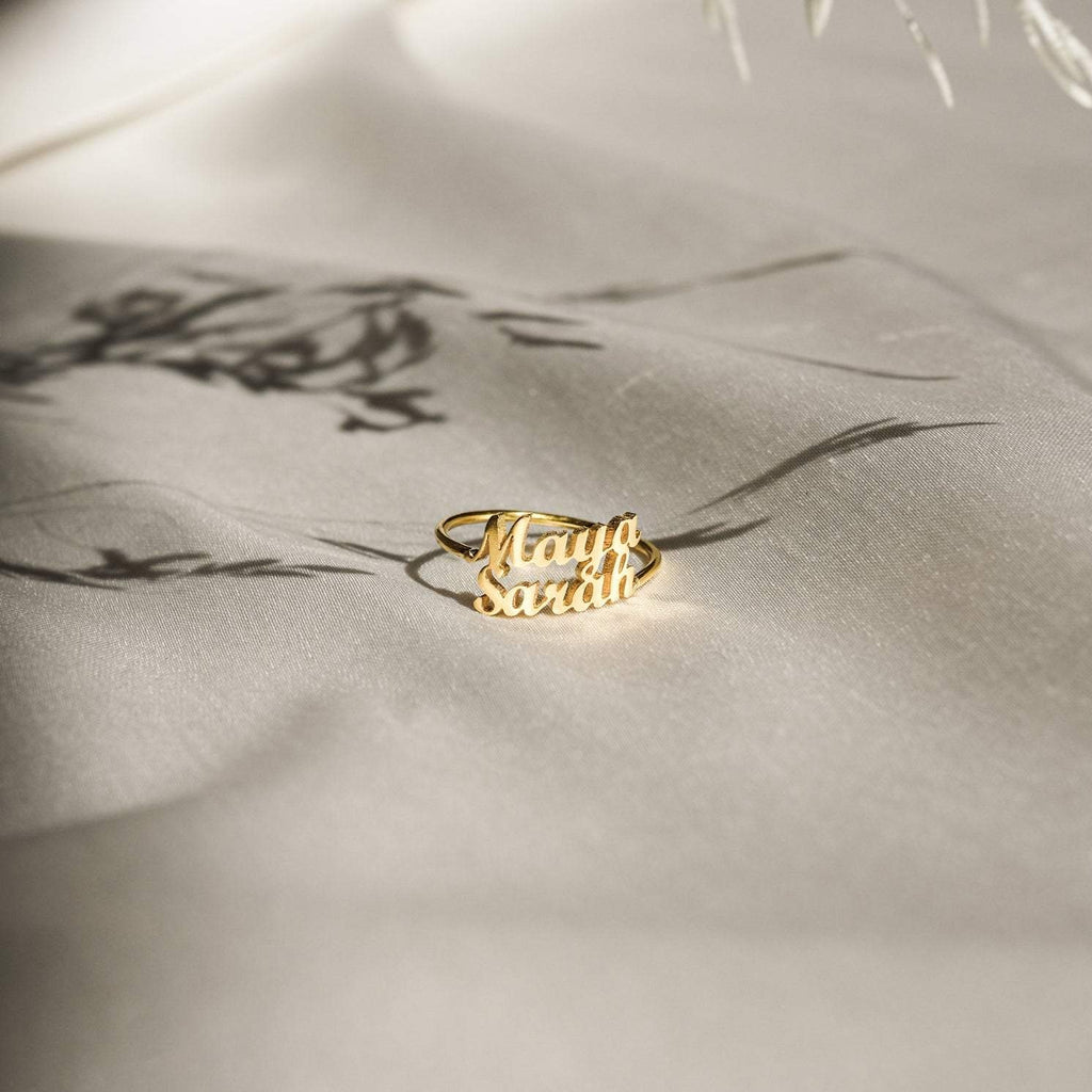 The London Duo Name Ring, featuring Maya and Sarah in elegant script, rests on soft cream fabric—a stunning custom jewelry piece that celebrates your unique bond.
