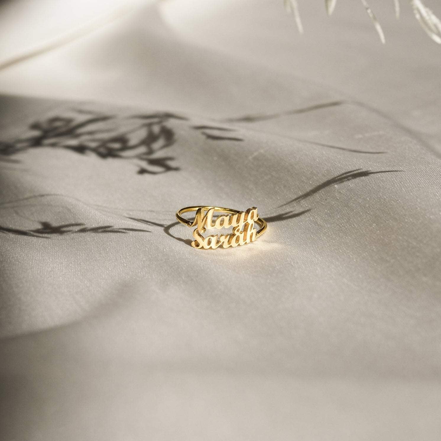 The London Duo Name Ring, featuring Maya and Sarah in elegant script, rests on soft cream fabric—a stunning custom jewelry piece that celebrates your unique bond.