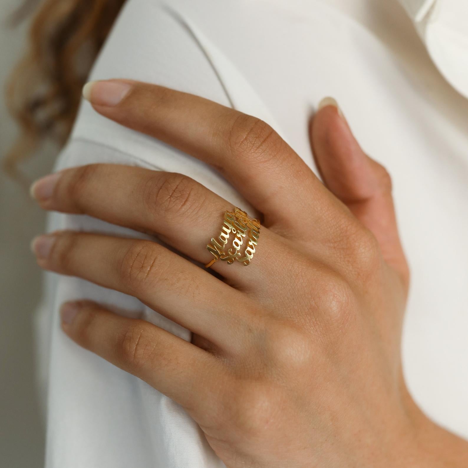 A hand with neatly manicured nails wears the London Triple Name Ring in gold, engraved with custom names, resting on a white shirt—ideal for fans of personalized jewelry.