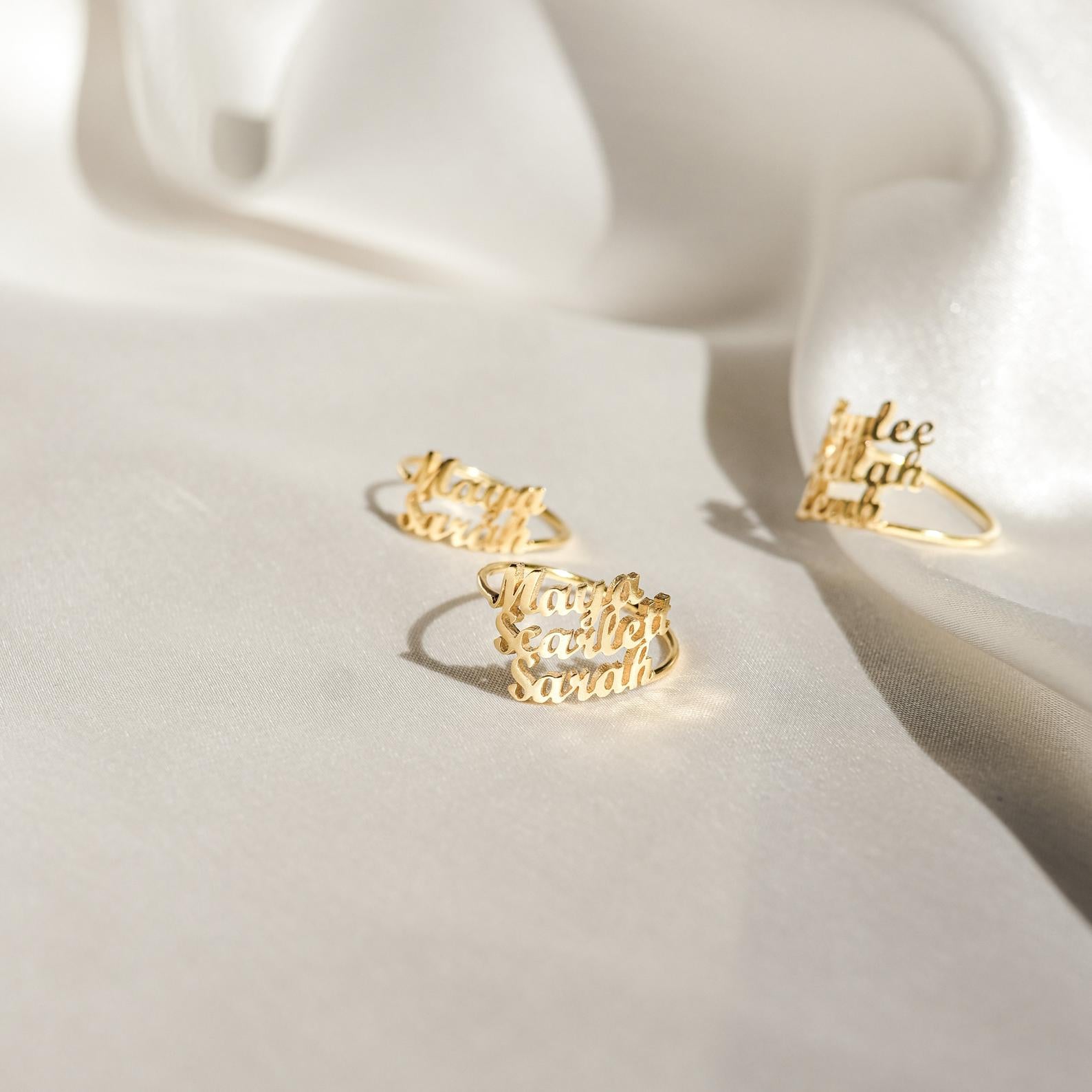 Three London Triple Name Rings with different engraved names rest on soft, white satin fabric, highlighting the elegance of personalized jewelry.