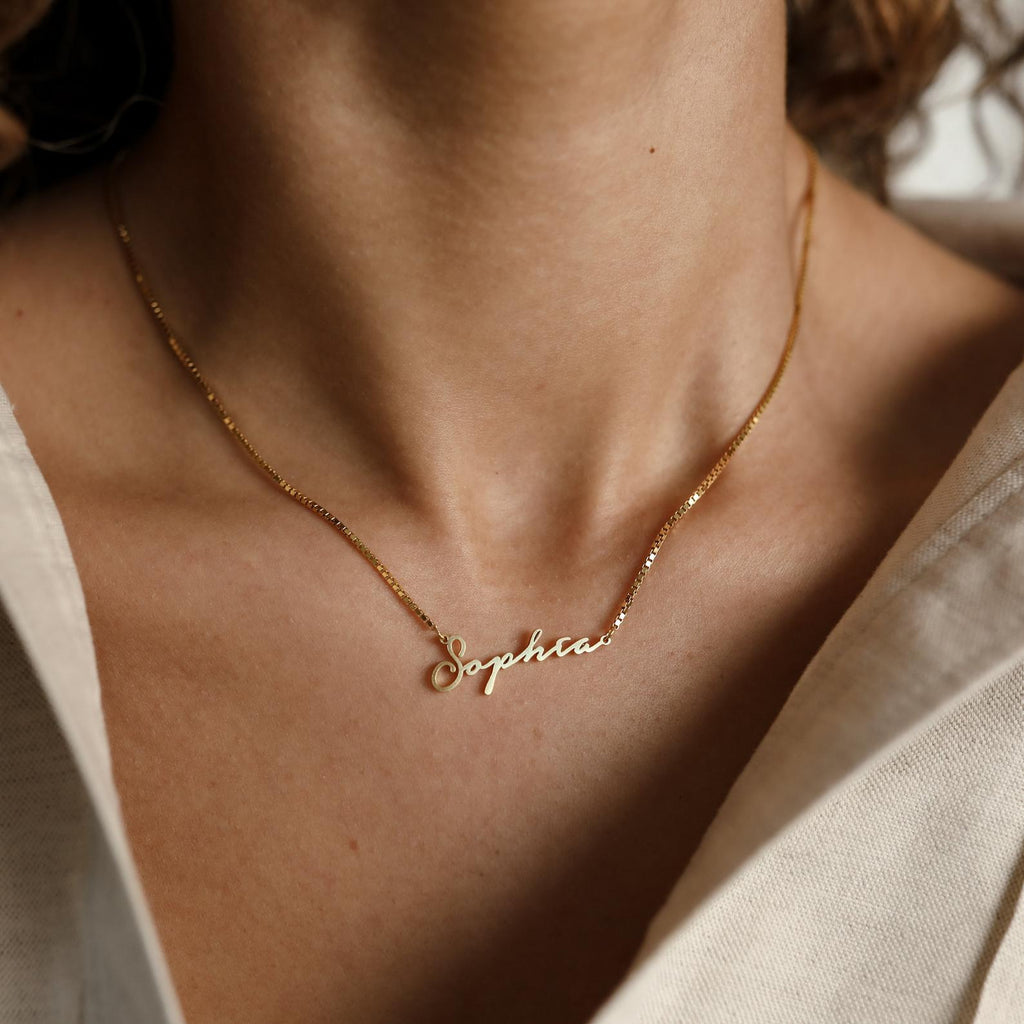 The Venice Name Necklace in Box Chain, customized with the name Sophia, shines elegantly on a wearer dressed in a cream-colored shirt.