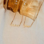 Two modern Mellow Name Necklaces in Box Chain, personalized with “Aubrey” and “Maya,” sit beside an amber glass vase. One showcases a sleek box chain for a contemporary touch.