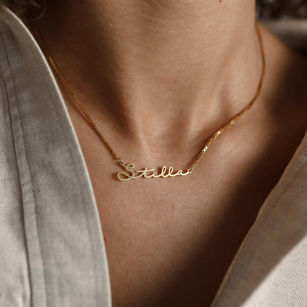 A gold Summer Name Necklace in box chain, featuring the name Stella in cursive, rests on a person’s neck with a light shirt—an elegant piece perfect for adding a personalized touch to your look.