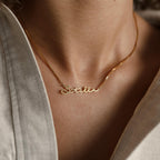 A gold Summer Name Necklace in box chain, featuring the name Stella in cursive, rests on a person’s neck with a light shirt—an elegant piece perfect for adding a personalized touch to your look.