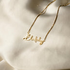 Elegant jewelry at its finest: the Summer Name Necklace in Box Chain showcases "Blakely" in graceful cursive, beautifully set against a soft cream-colored fabric background.