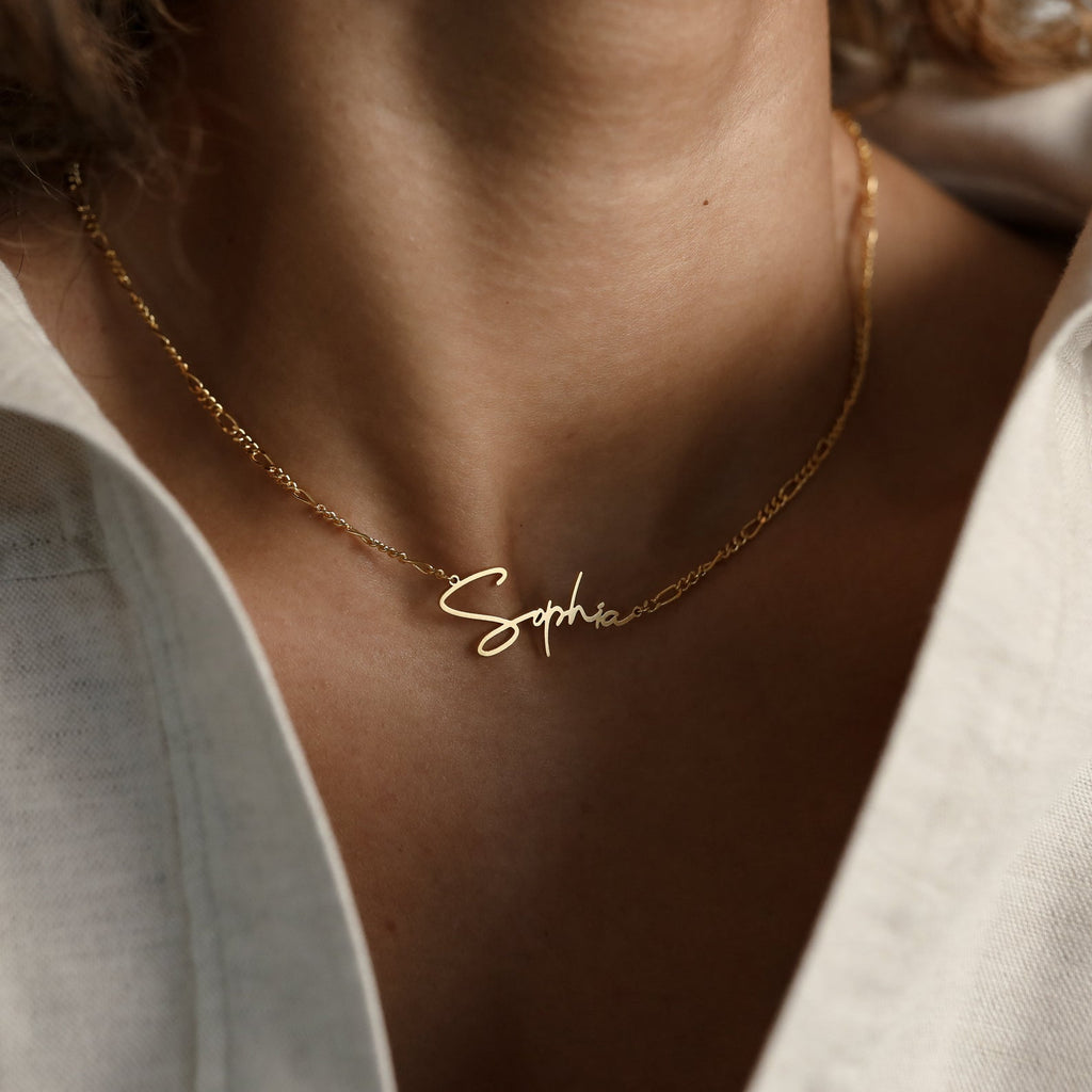 The Summer Name Necklace in Figaro Chain features a cursive nameplate spelling "Sophia," elegantly draped around a woman's neck—the perfect personalized accessory for summer.