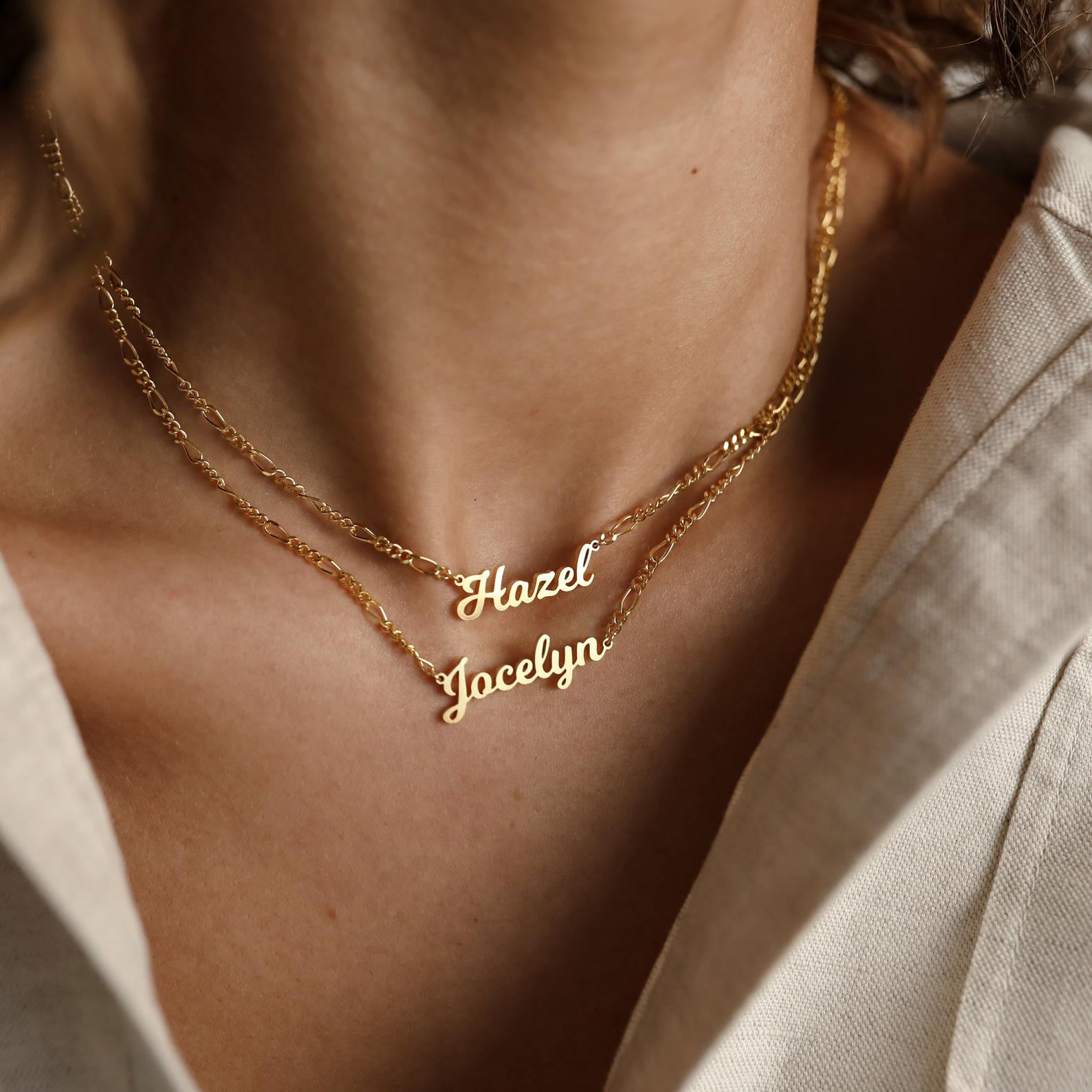 Two Mellow Name Necklaces in Figaro Chain, personalized with "Hazel" and "Jocelyn," are worn together. Each gold necklace features a sleek Figaro chain for an elegant touch.