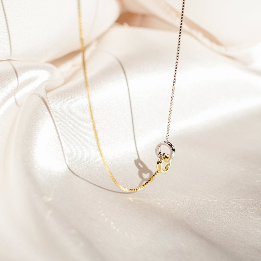 Two-tone eternity circle necklace with interlocked gold and silver rings, displayed on soft ivory satin, highlighting its delicate symbolism and contrast in metals.