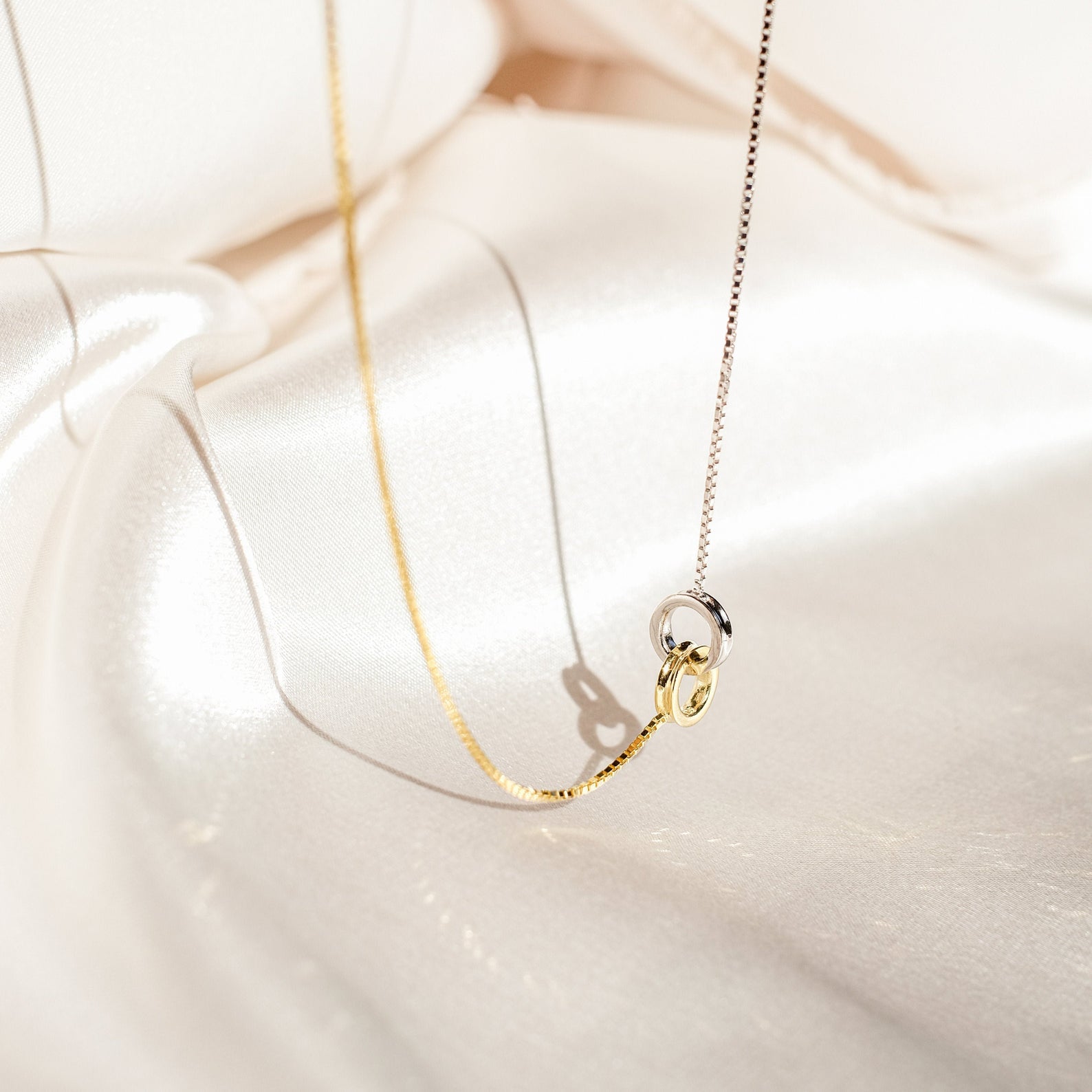 Two-tone eternity circle necklace with interlocked gold and silver rings, displayed on soft ivory satin, highlighting its delicate symbolism and contrast in metals.