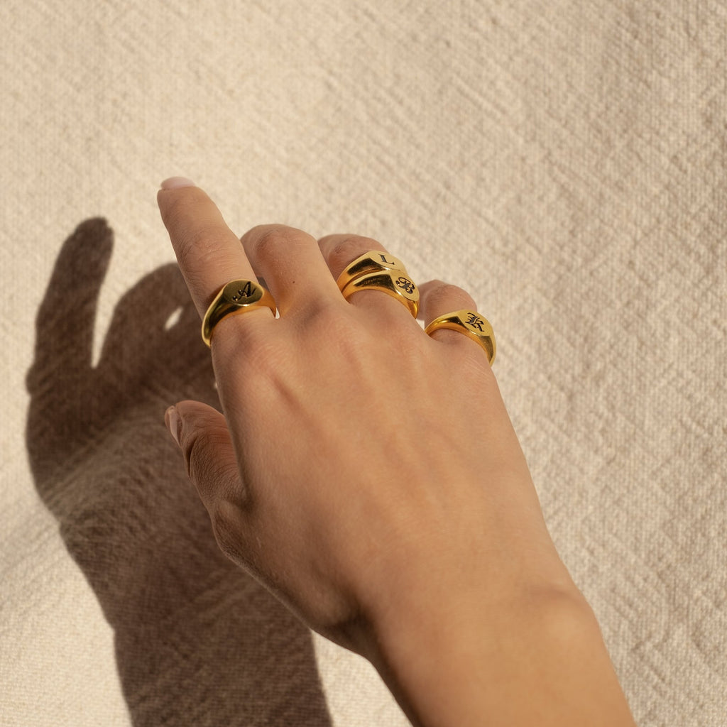 A hand wearing three gold rings, including the Initial Signet Ring, rests on textured beige fabric in sunlight.