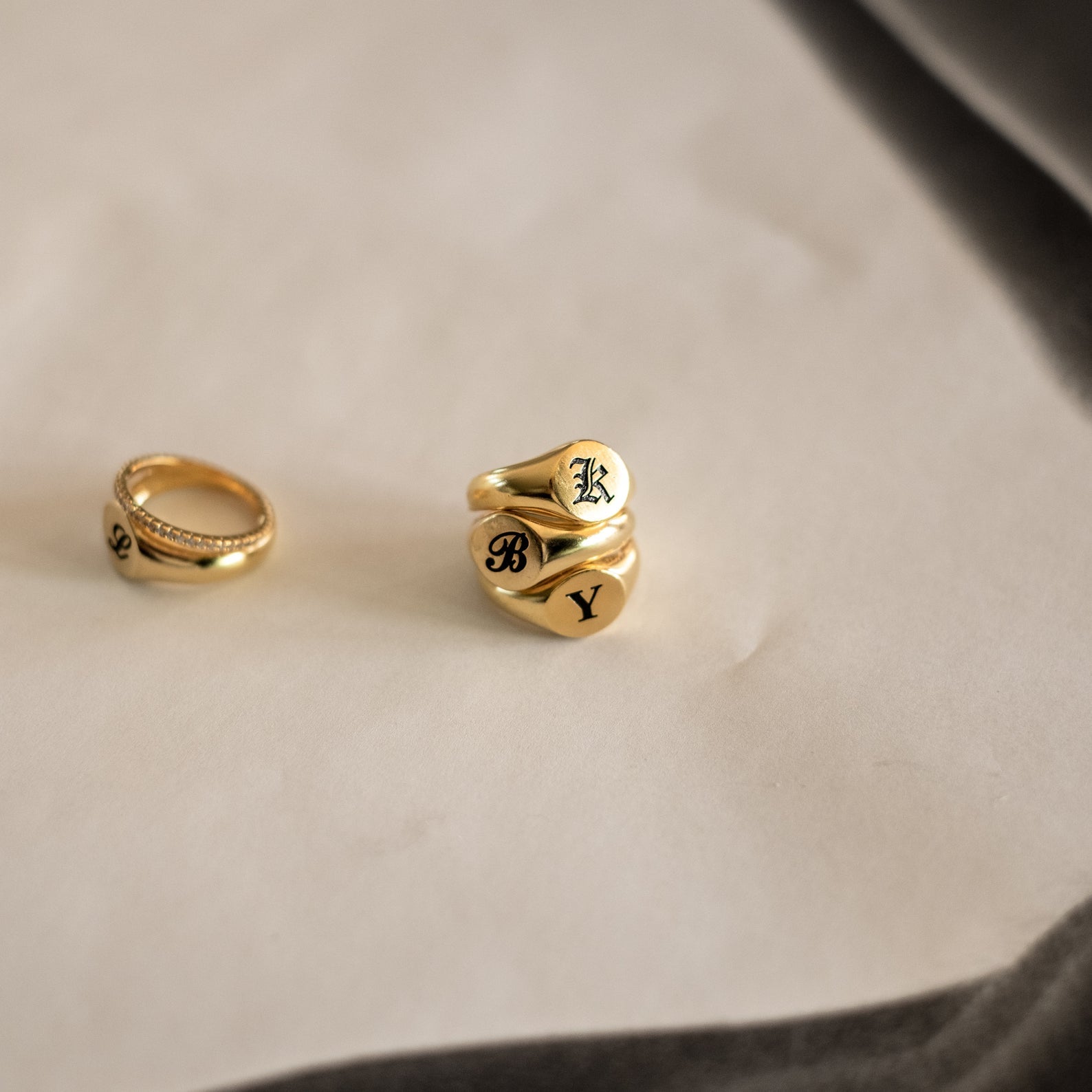 Four gothic Initial Signet Rings rest on a light surface—three engraved with K, B, and Y, and one customized ring adorned with small stones.