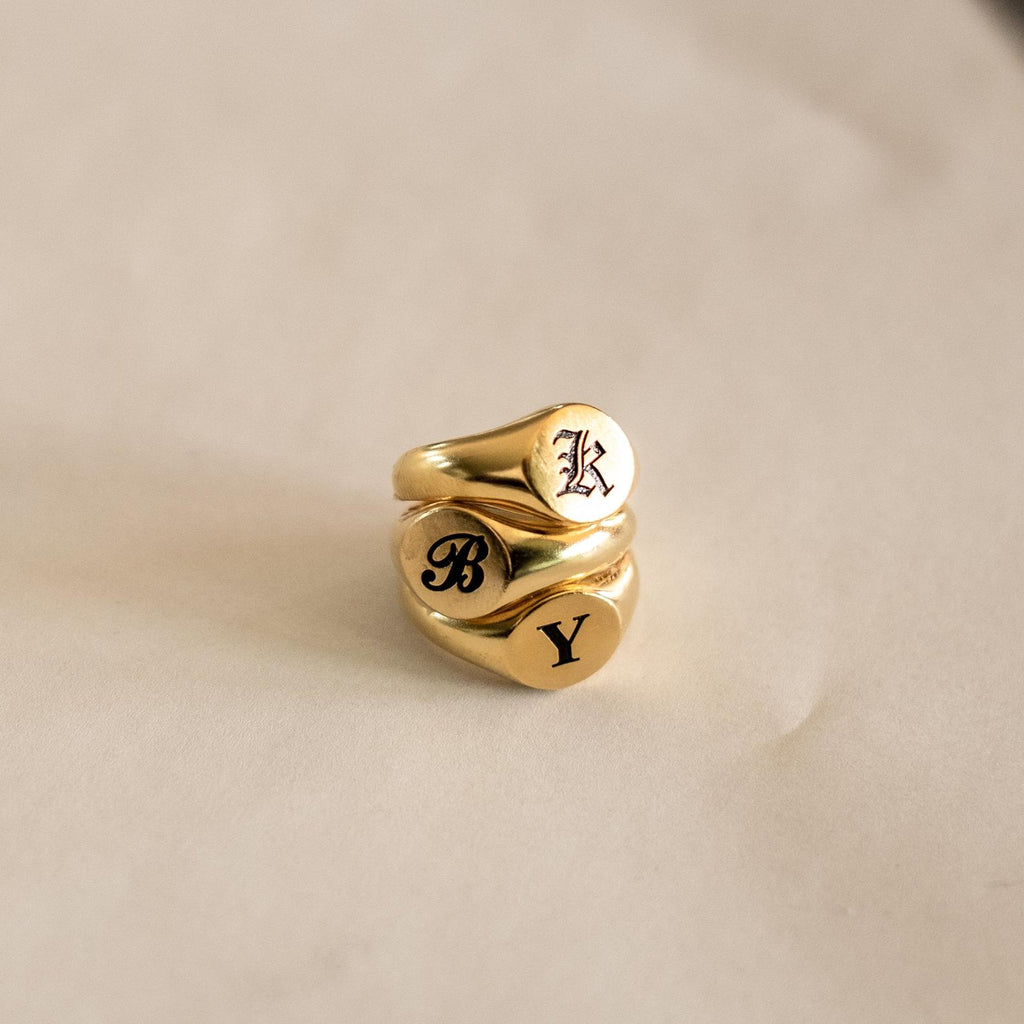 Three Initial Signet Rings in gold with gothic-style K, B, and Y engravings are stacked on a light beige surface.