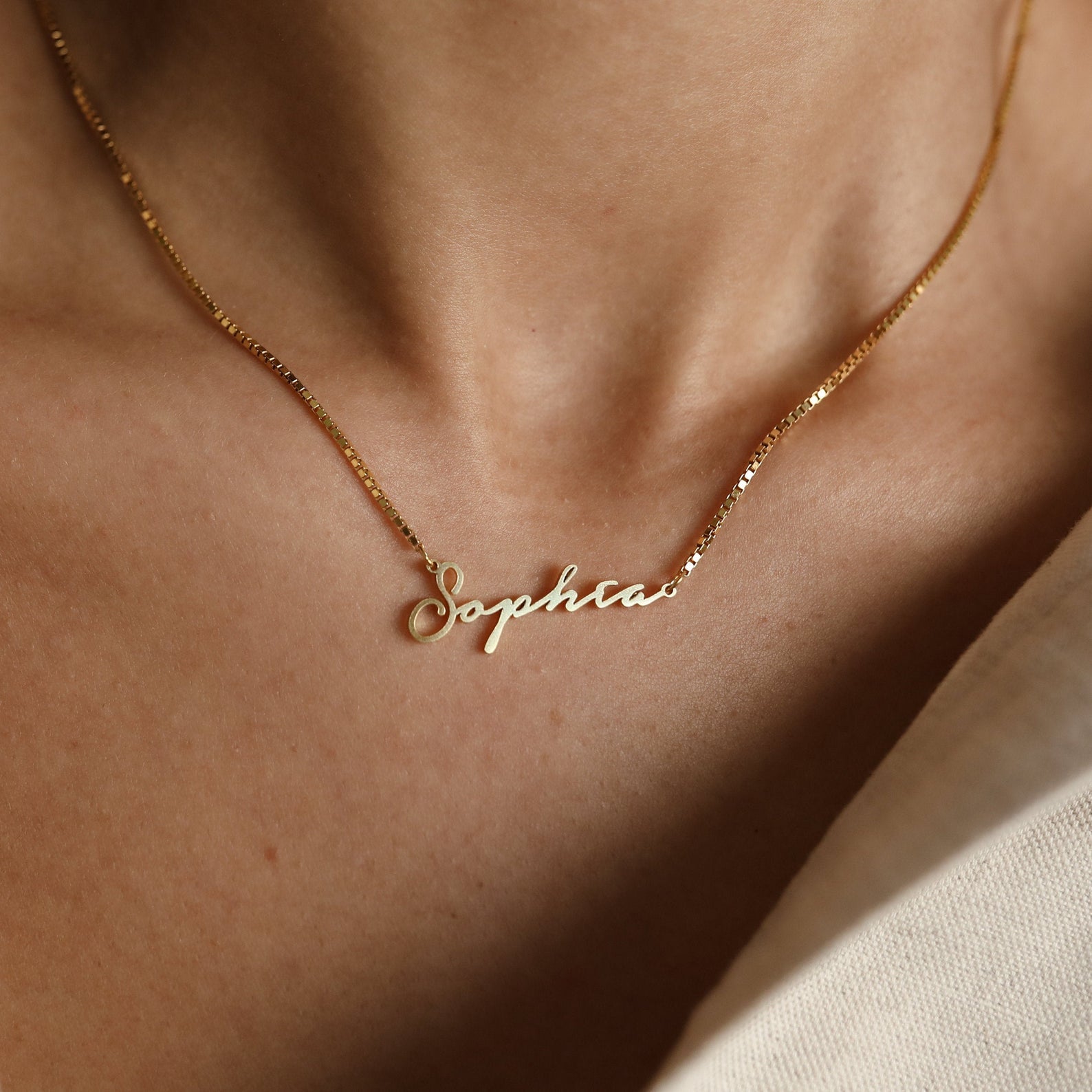 The Venice Name Necklace in Box Chain gleams on a person's neck, its gold shine standing out beautifully against a cream-colored garment.