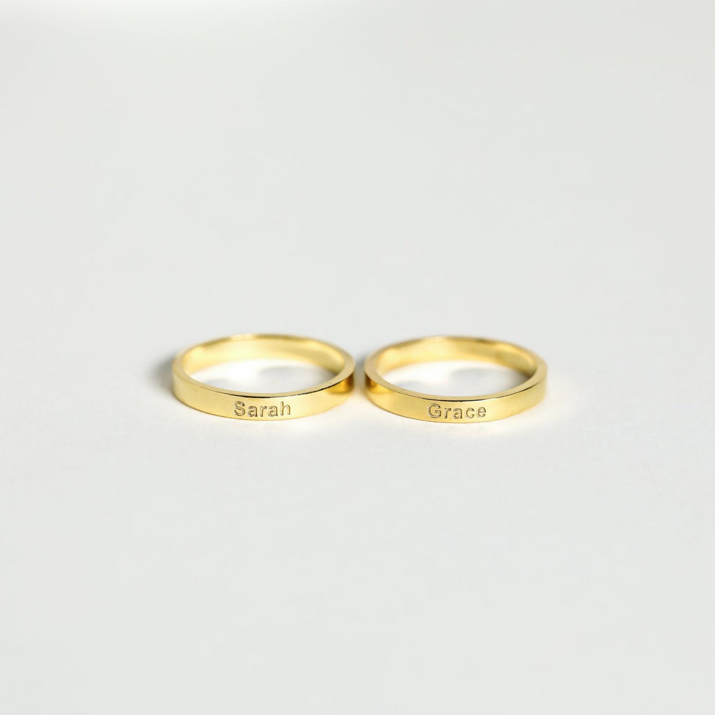 Two Alice Thin Engraved Rings rest on a white surface, each customized with names—a perfect personalized gift.