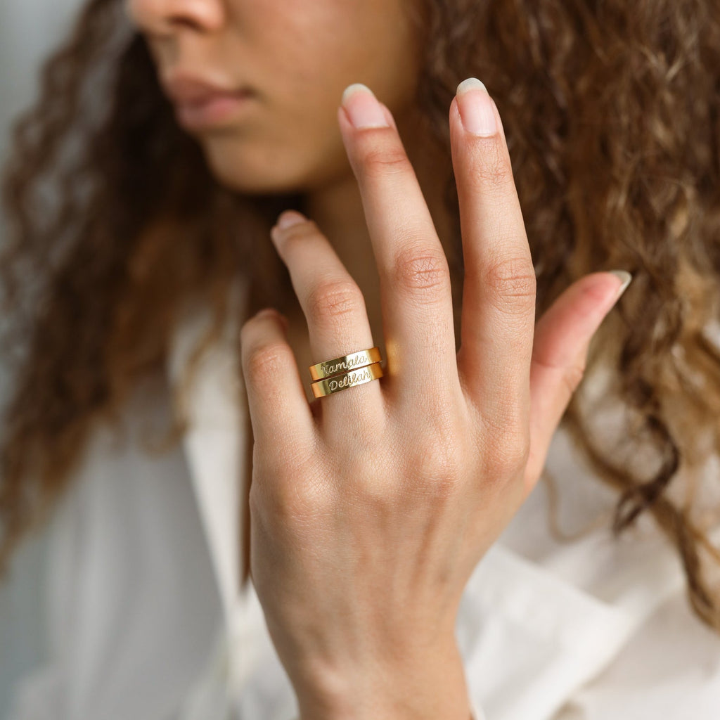 A woman with curly hair wears two stylish Ann Engraved Name Rings on her middle finger, featuring the engravings “Family” and “Dad.”.