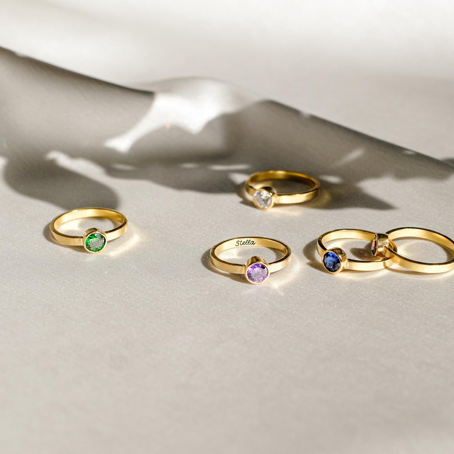 A Birthstone Signet Ring and four other gold rings rest on a soft, cream-colored surface with gentle shadows.