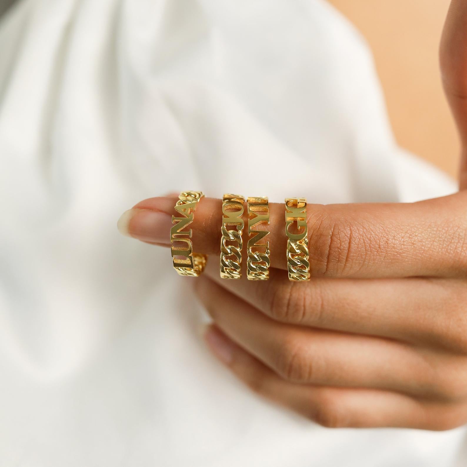 A hand wears three Newsletter Curb Name Rings in Rose Gold on one finger, set against a soft white fabric background.