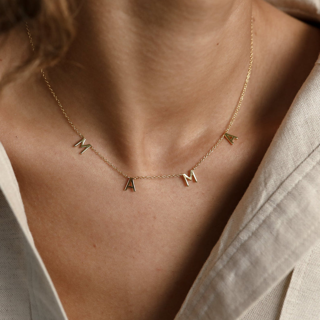 Gold “MAMA” letter necklace worn at the collarbone, featuring four spaced capital letter charms on a delicate chain, styled against a neutral-toned outfit.