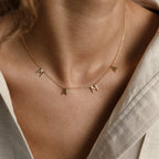 Gold “MAMA” letter necklace worn at the collarbone, featuring four spaced capital letter charms on a delicate chain, styled against a neutral-toned outfit.