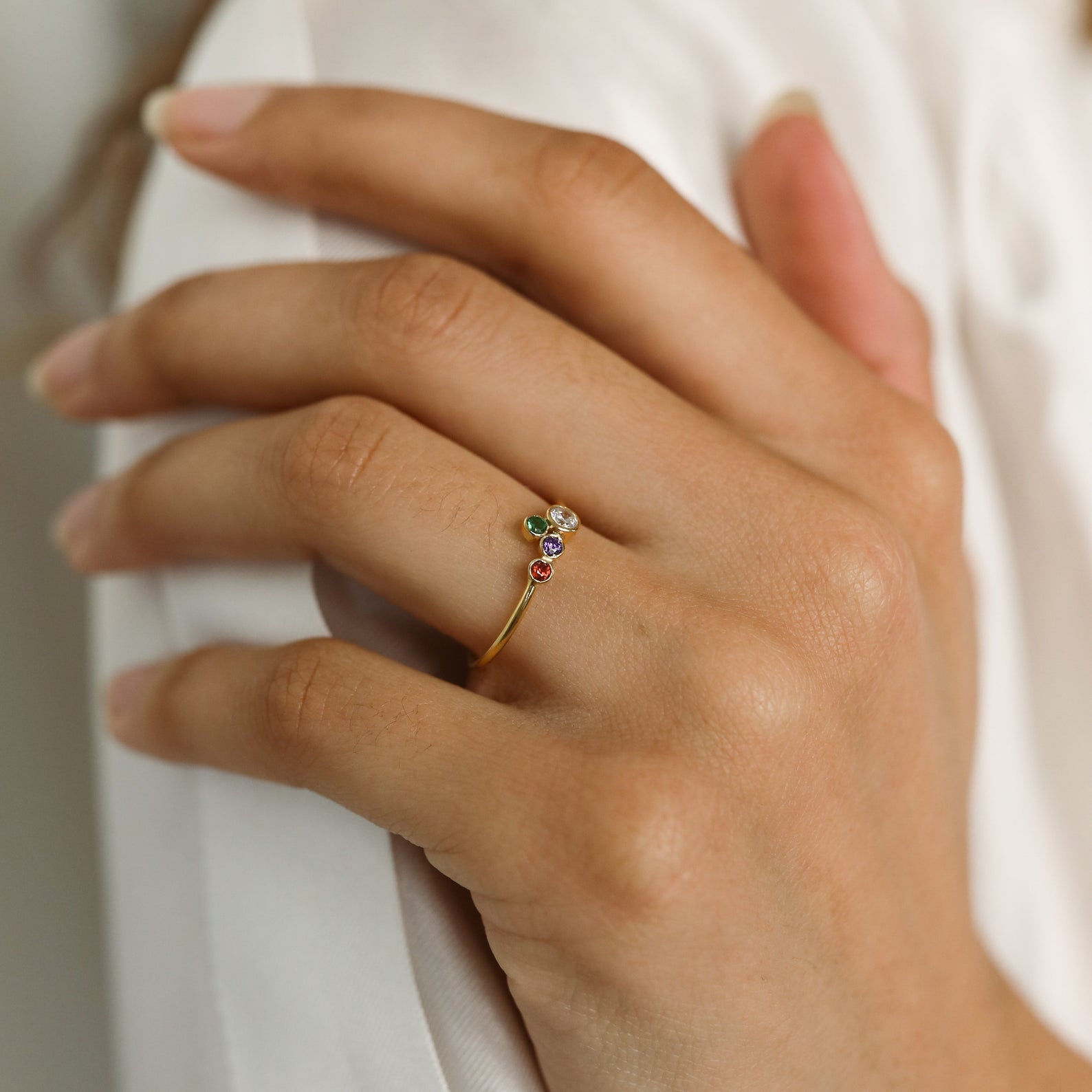 A hand displays the Cluster Birthstone Ring—a thin gold band with three colorful gemstones on the ring finger, featuring a classic design.