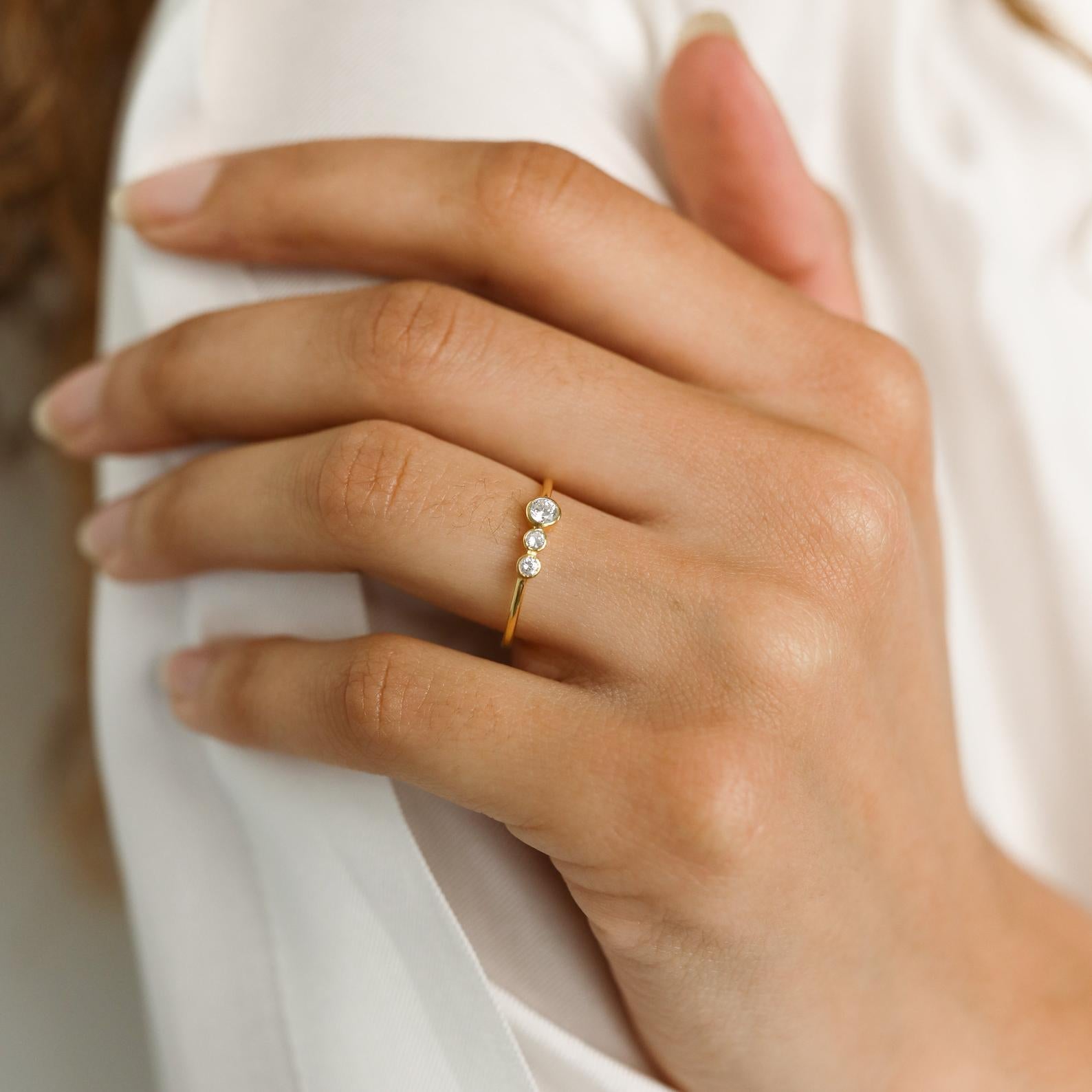 A hand wearing the Cluster Birthstone Ring, featuring three small diamonds on a thin gold band, rests on white fabric—a subtle nod to classic design.