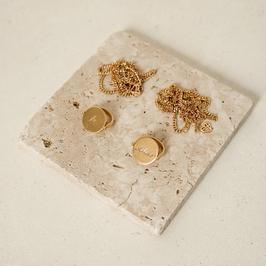 Two Spinner Pendant Necklaces featuring engraved, round spinning pendants rest on a square stone tile against a light background.