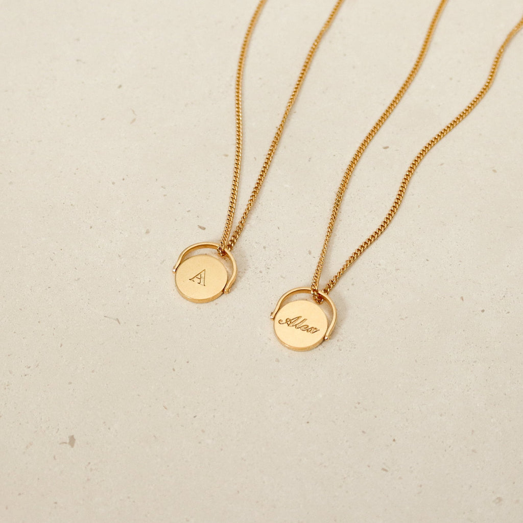Two Spinner Pendant Necklaces in gold, each with a round spinning detail pendant—one engraved with "A" and the other with "Allan"—are displayed on a beige surface.
