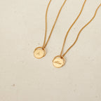 Two Spinner Pendant Necklaces in gold, each with a round spinning detail pendant—one engraved with "A" and the other with "Allan"—are displayed on a beige surface.