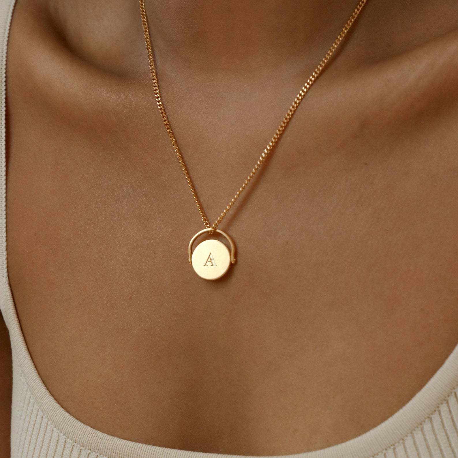 Spinner Pendant Necklace with a round pendant engraved with the letter A, shown on a model in a beige top. The spinning feature adds an interactive touch to this elegant accessory.