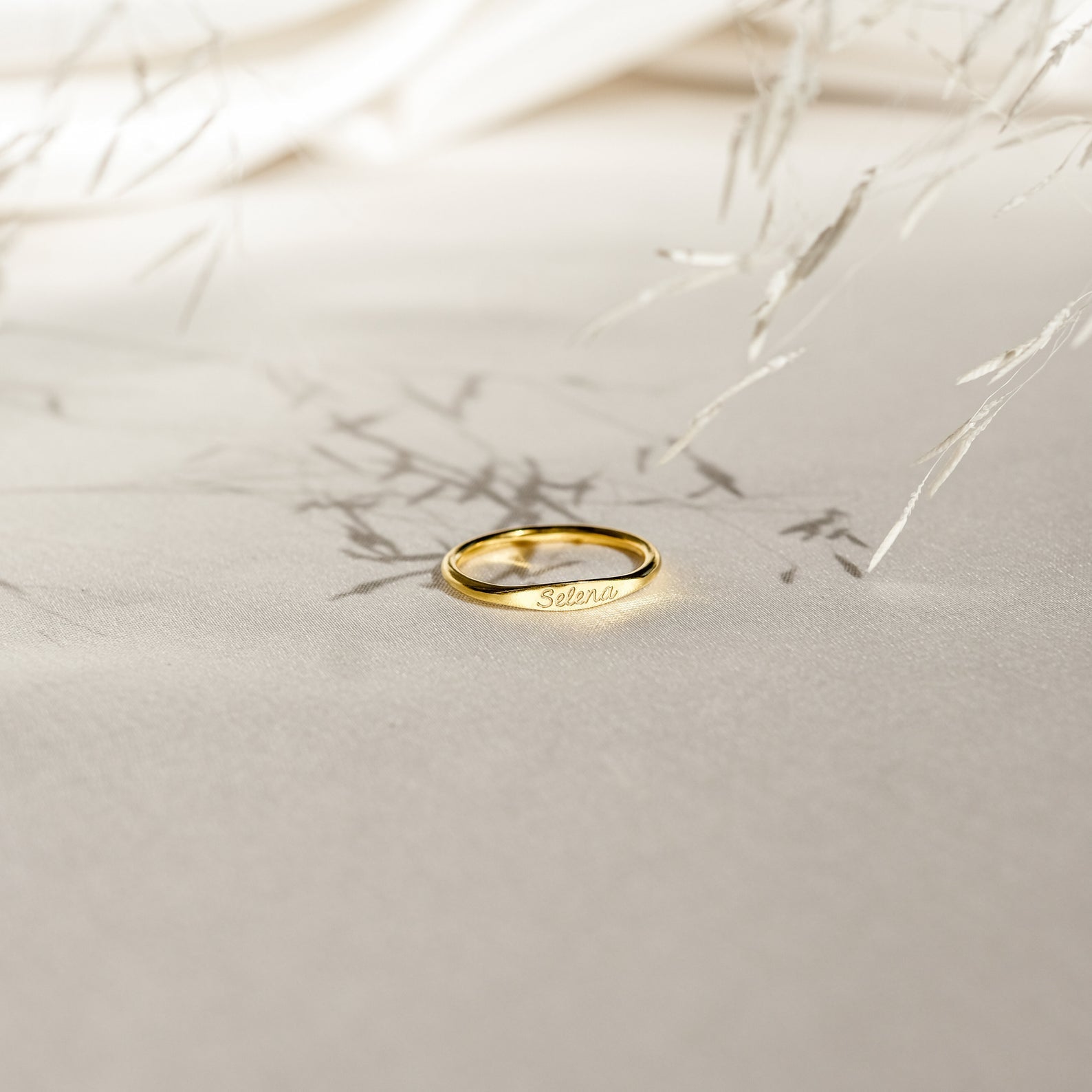 The Ann Skinny Name Ring in Rose Gold, engraved with "Selena," rests on a light beige surface with delicate plant shadows—a perfect Christmas gift for her.