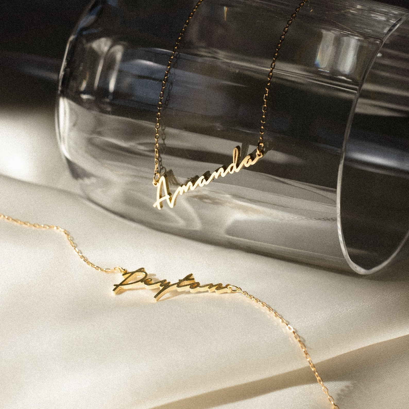 Two Venice Name Necklaces, "Amanda" and "Peyton," in elegant script, displayed on glass and satin. These dainty gold necklaces are perfect personalized jewelry pieces to elevate any look.