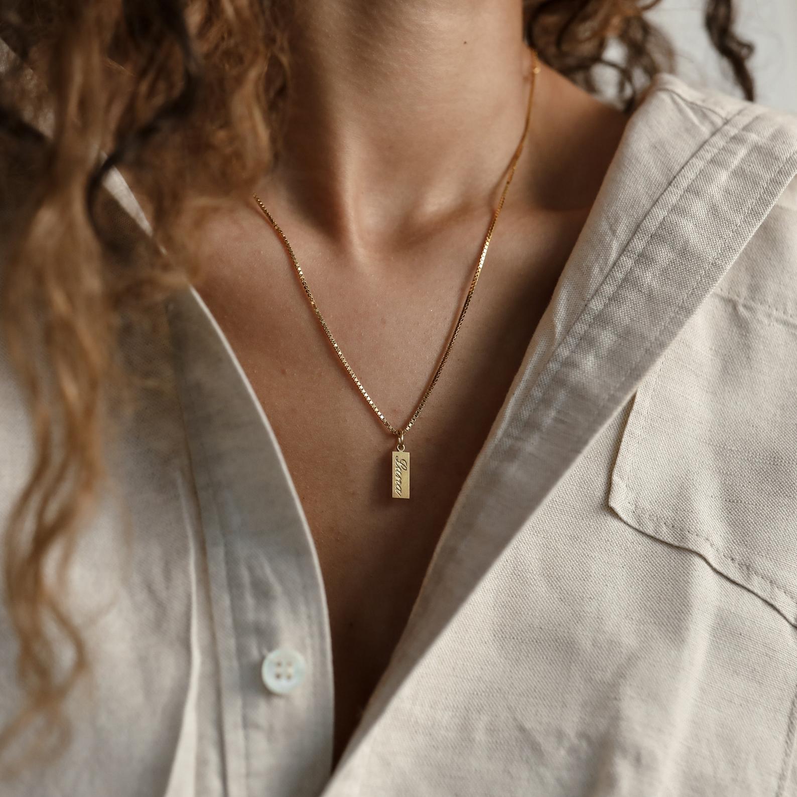 A woman wearing a cream shirt and the Dainty Tag Name Necklace in Rose Gold, featuring a rectangular pendant on a delicate chain.