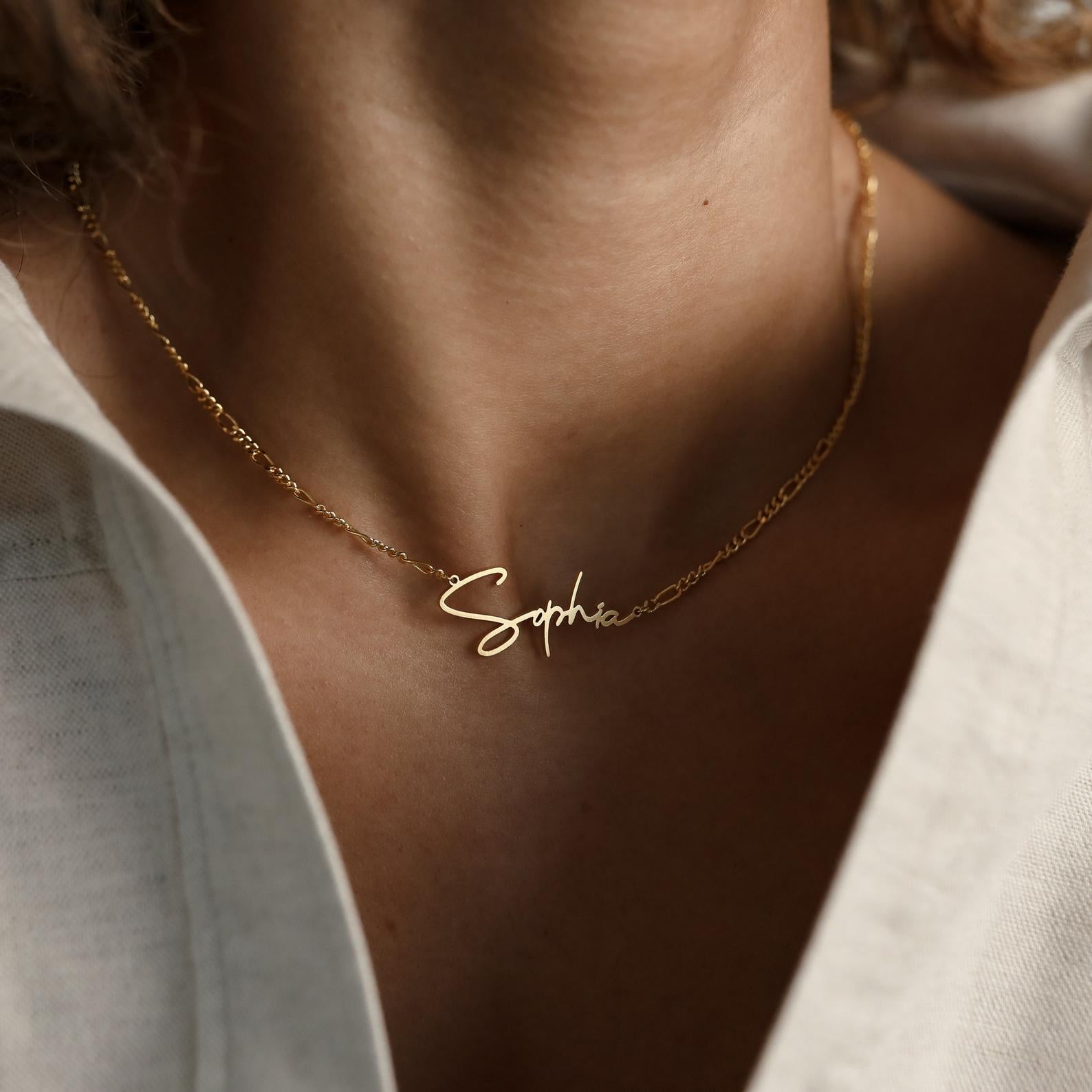 Minimal style Carrie Name Necklace in Figaro Chain, gold finish with "Sophia" nameplate, worn elegantly over a white shirt with an open collar.