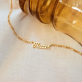 The Mellow Name Necklace in Figaro Chain with a Hazel pendant is displayed beside a glass object on cream fabric.
