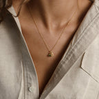 A person wears a beige shirt and the Zodiac Birthstone Necklace, a gold necklace with small charms, shown from neck to chest.