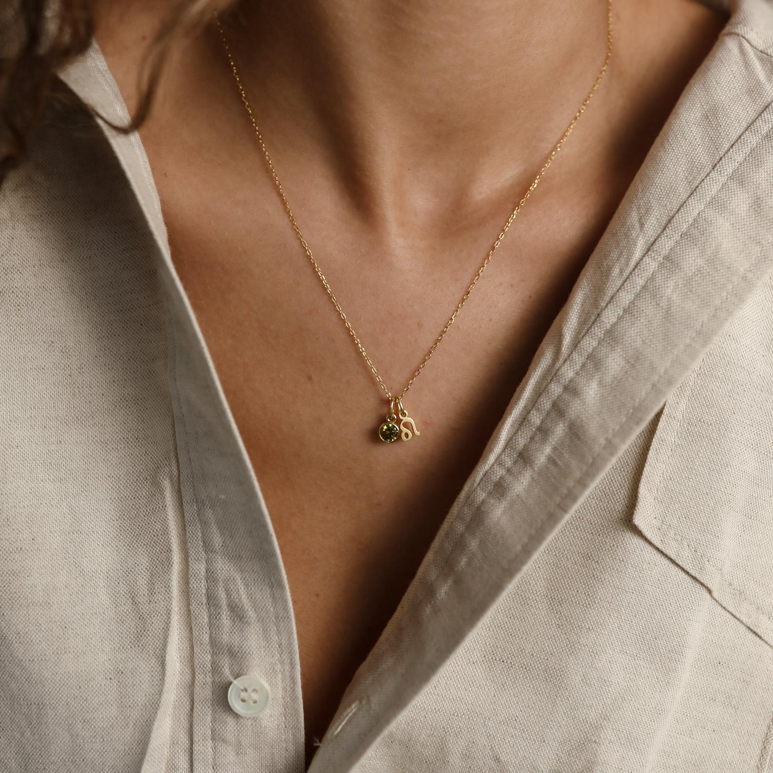 A person wears a beige shirt and the Zodiac Birthstone Necklace, a gold necklace with small charms, shown from neck to chest.